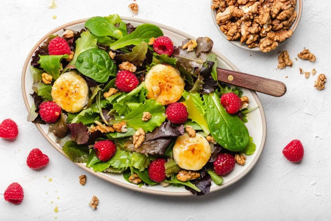 Salat mit gebackenem Ziegenkäse mit Honig, Walnüssen und frischen Himbeeren angerichtet auf einem Teller.