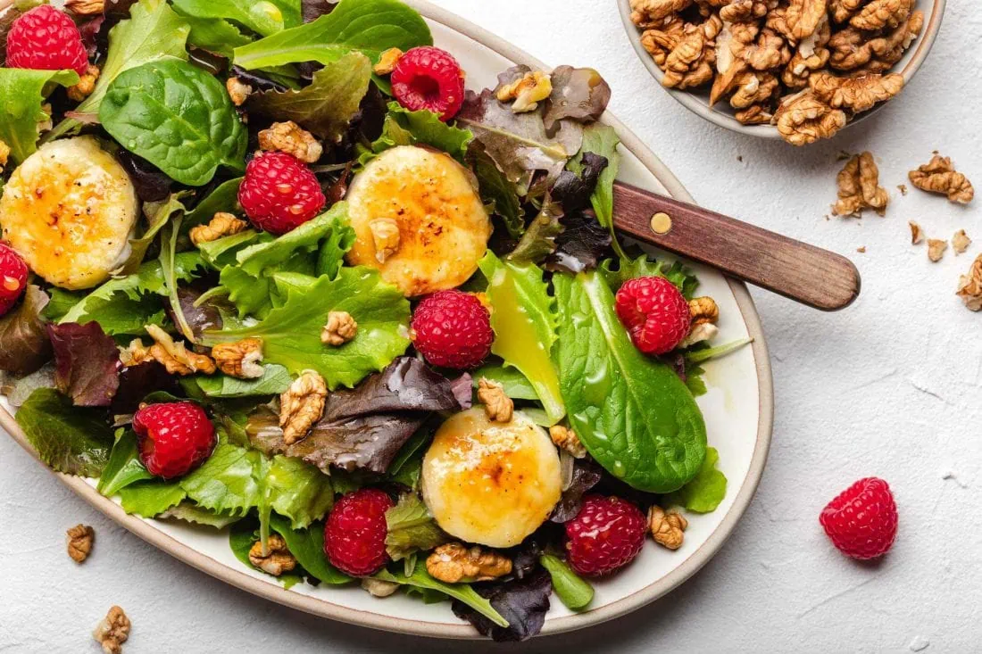 Bunter Blattsalat mit karamellisiertem Ziegenkäse serviert auf einem Teller mit Himbeeren, Walnüssen und einer Vinaigrette.