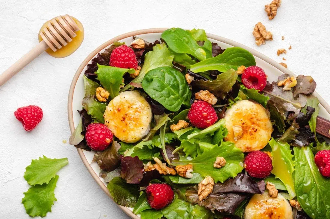 Salat mit Ziegenkäse und Honig angerichtet mit Himbeeren und gehackten Walnusskernen auf einem ovalen Teller.