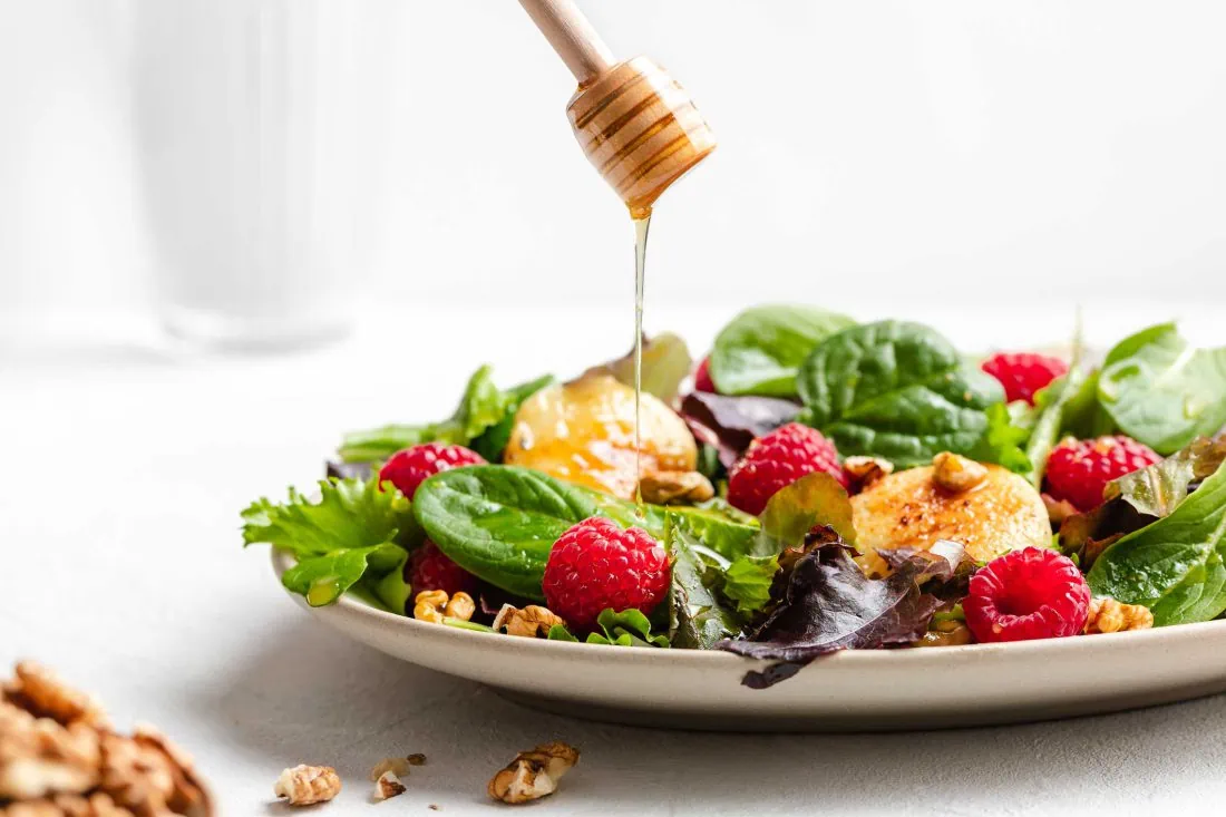 Salat mit gebackenem Ziegenkäse mit Honig und Walnüssen wird mit einem Honiglöffel beträufelt.