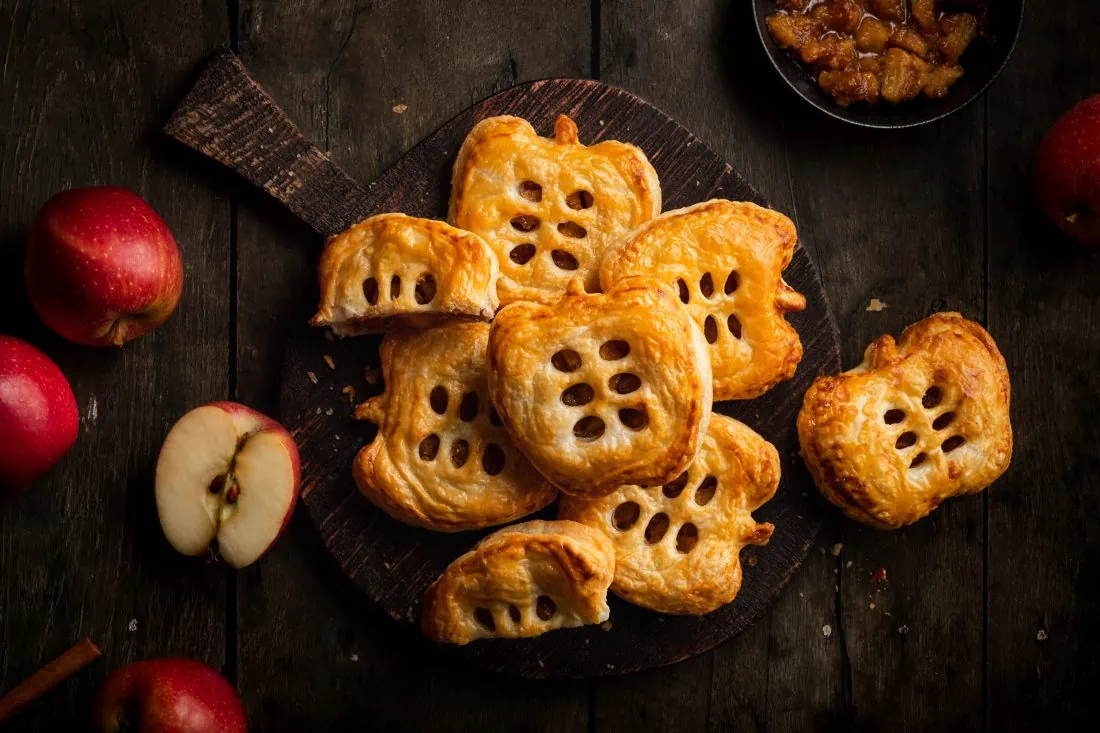 Schnelle Apfeltaschen mit Blätterteig serviert auf einem Holzbrett. Daneben liegen mehrere Äpfel und eine Schale mit der Füllung.