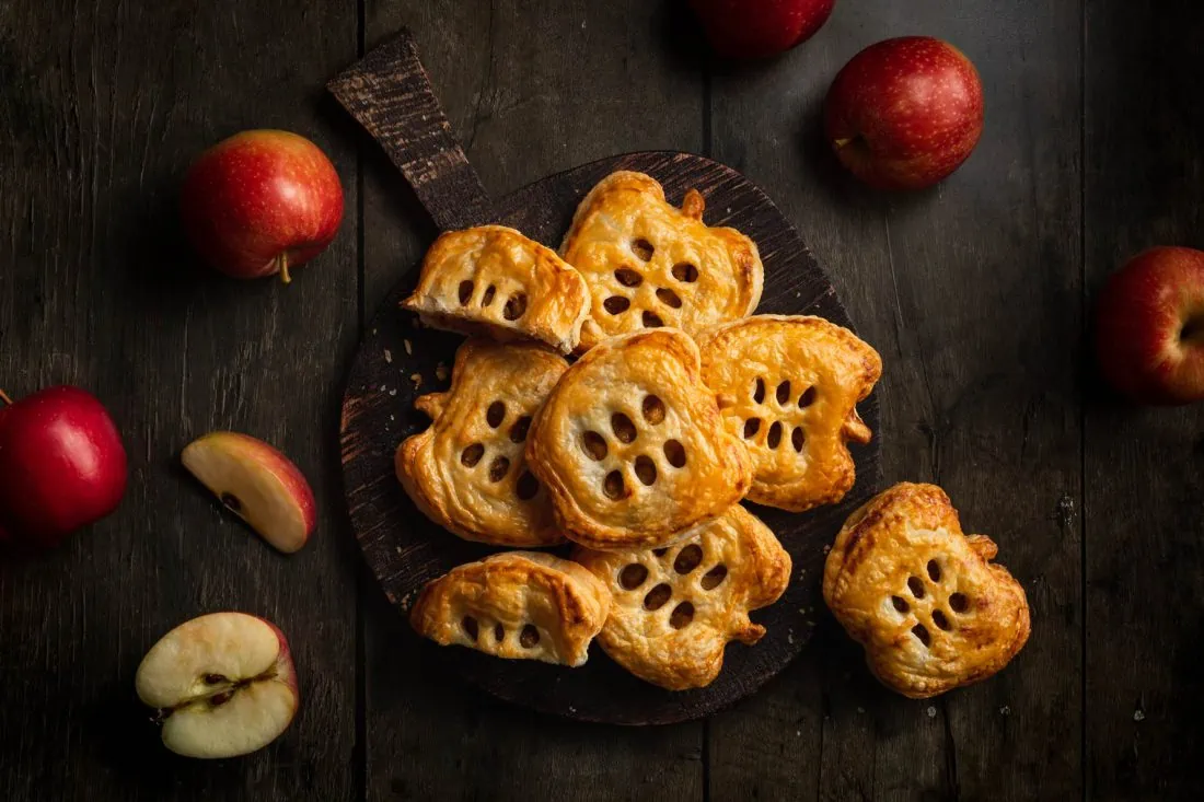 Schnelle Apfeltaschen mit Blätterteig in passender Apfelform auf einem Holzbrett. Daneben liegen mehrere Äpfel.