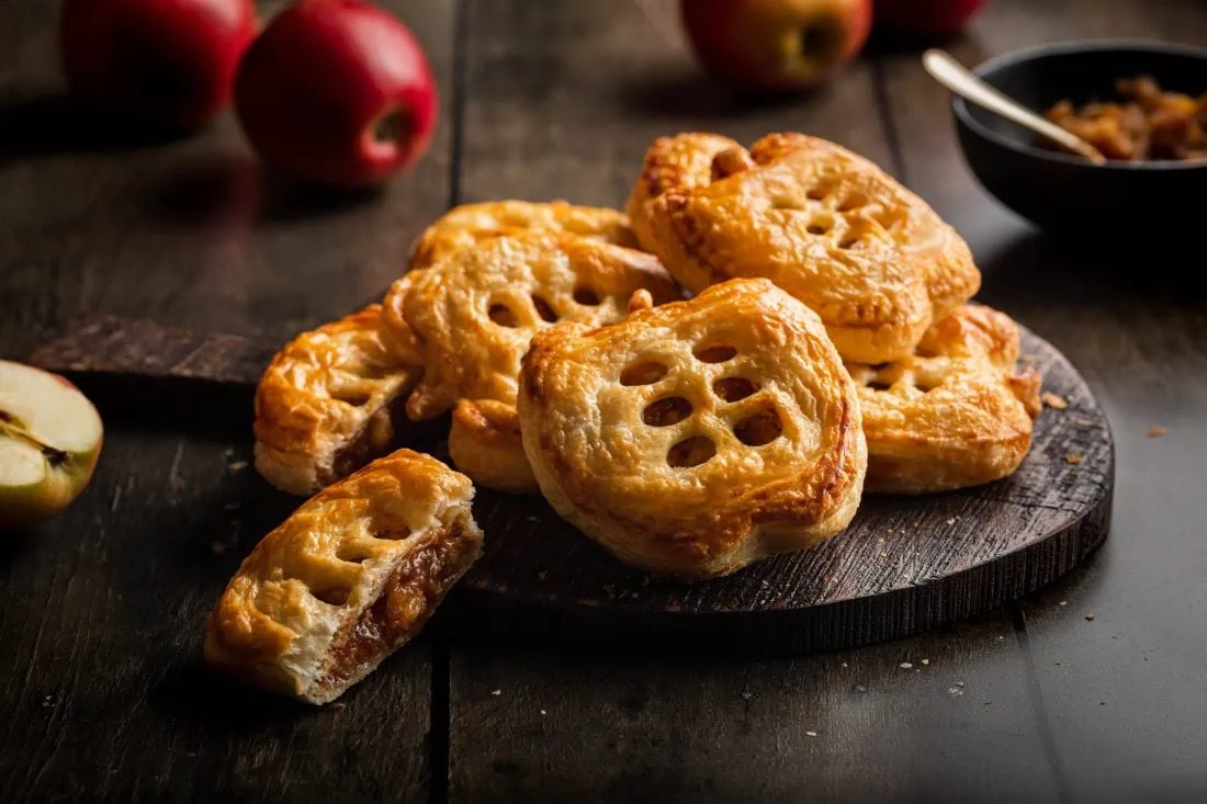 Mehrere Blätterteigtaschen mit einer zimtigen Apfelfüllung auf einem Holzbrett. Eine Apfeltasche liegt angeschnitten daaneben.