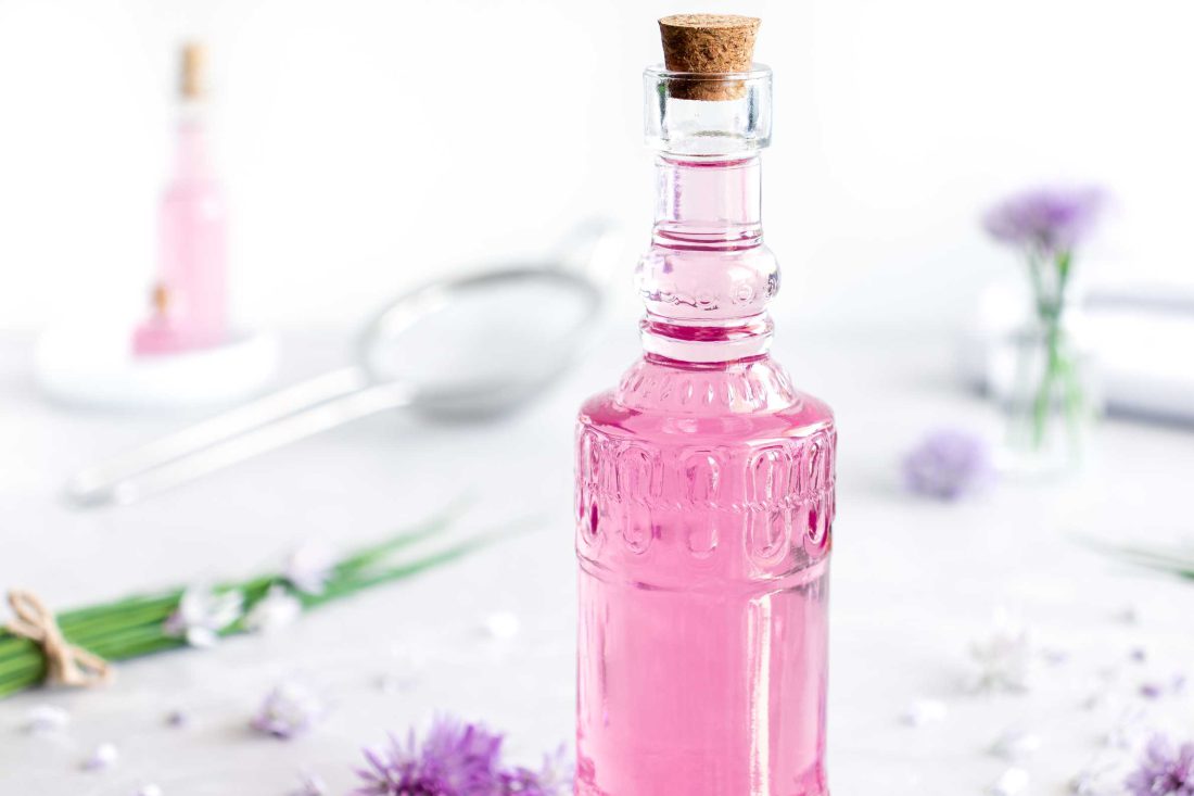 Schnittlauchblütenessig in einer Flasche mit Korken. Neben dem rosa Schnittlauchblütenessig liegen mehrere lila Schnittlauchblüten.