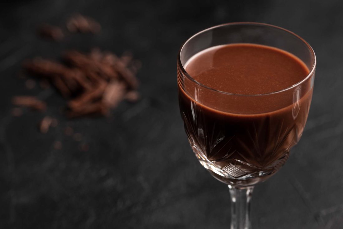 Schoko-Eierlikör in einem Likörglas auf einem dunklen Untergrund. Neben dem Glas liegt geschnittene Schokolade.