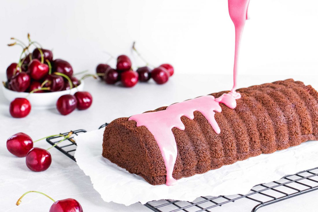Schoko-Kirsch-Kuchen, der mit einer rosa Glasur aus Zuckerguss überzogen wird. Im Hintergrund liegen frische Kirschen.