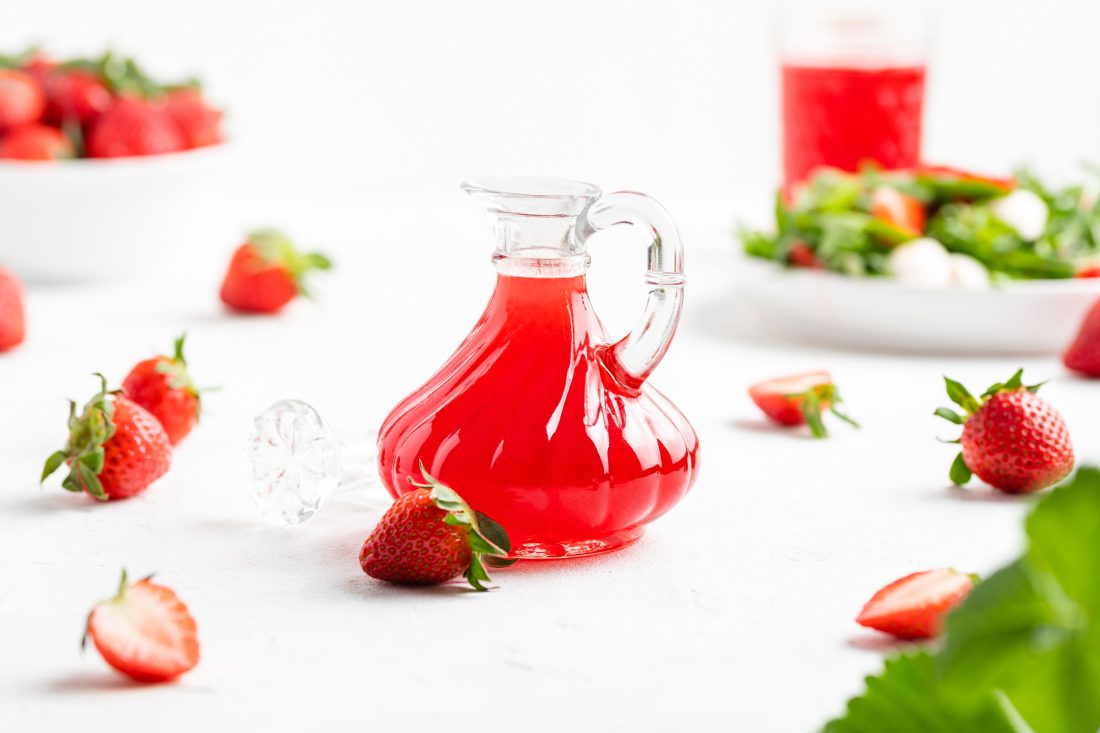 Der fruchtige, rote Essig abgefüllt in einer schönen Flasche. Daneben liegen halbierte und ganze Erdbeeren.