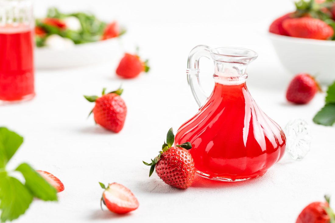 Der knallrote Fruchtessig abgefüllt in einer dekorativen Flasche. Neben dem Fruchtessig liegen frische Erdbeeren.