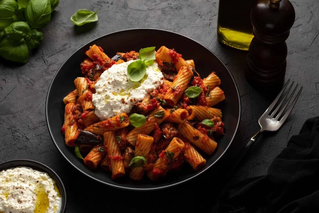 Sizilianische Pasta mit Ricotta und Auberginen in einem tiefen Teller. Neben dem Nudelgericht stehen eine Ölflasche und eine Pfeffermühle.