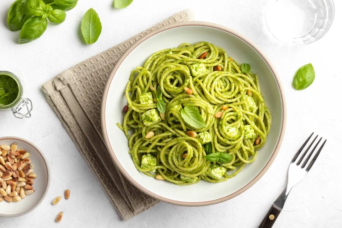 Spaghetti-Salat mit Pesto und Feta in einem Teller. Neben dem Salat liegen eine Gabel und die Zutaten.