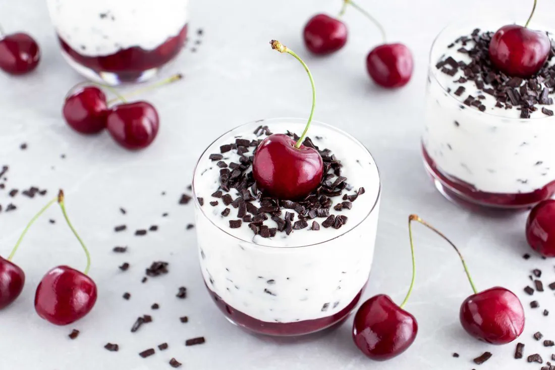 Stracciatella Creme mit Kirschen im Glas auf einem hellen Untergrund garniert mit frischen Kirschen und Schokoraspeln.