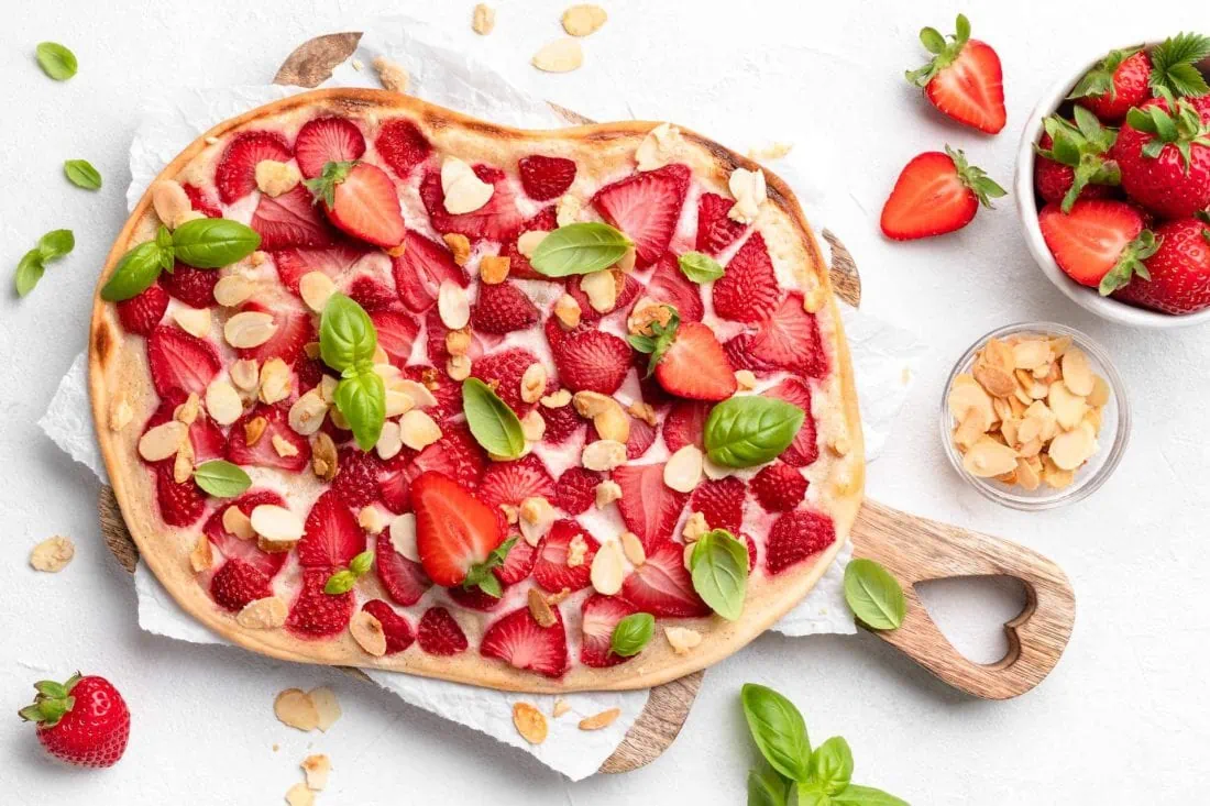 Süßer Flammkuchen mit Erdbeeren auf einem Holzbrett. Daneben stehen eine Schüssel mit karamellisierten Mandeln und frischen Erdbeeren.