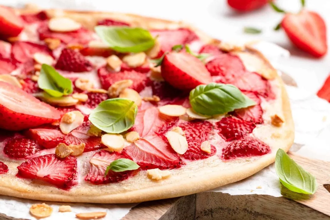 Süßer Erdbeer-Flammkuchen mit frischem Basilikum und gerösteten Mandelsplittern auf einem Holzbrett mit Backpapier.