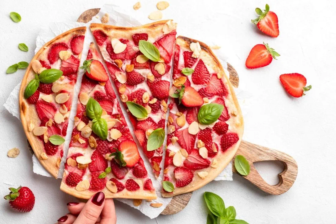 Süßer Flammkuchen mit Erdbeeren auf geschnitten auf einem Brett. Gerade nimmt sich jemand ein Stück davon.