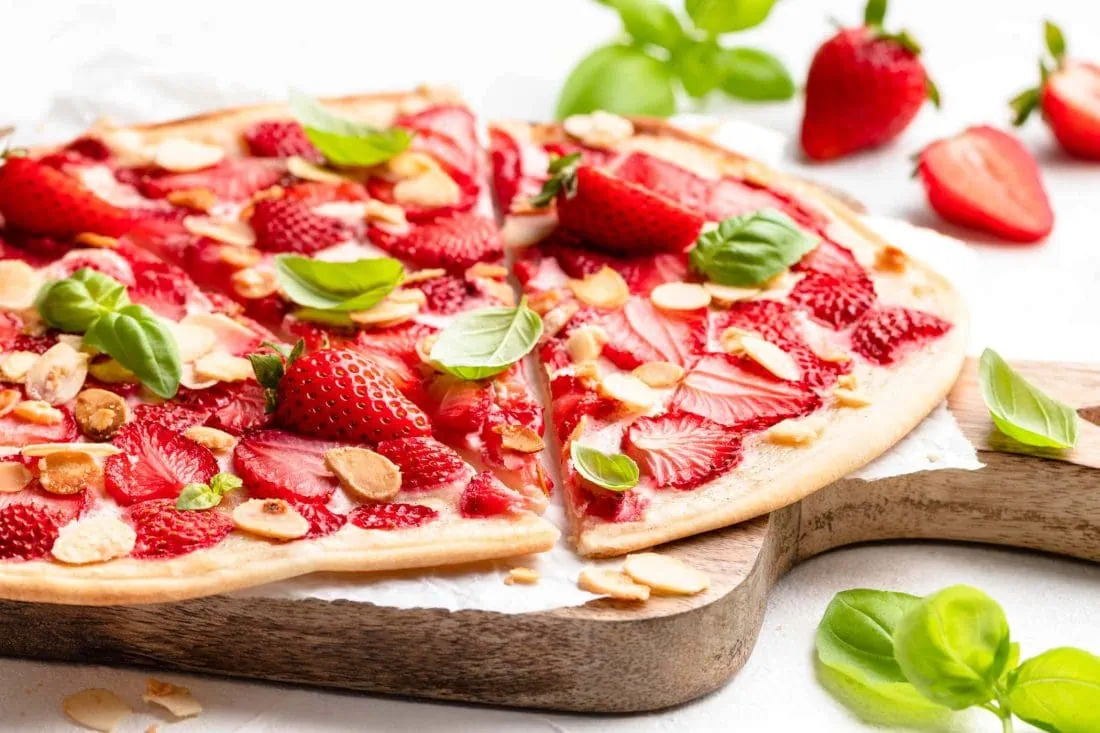 Süßer Flammkuchen mit frischen Erdbeeren, Basilikum und gerösteten Mandeln auf einem Holzbrett.