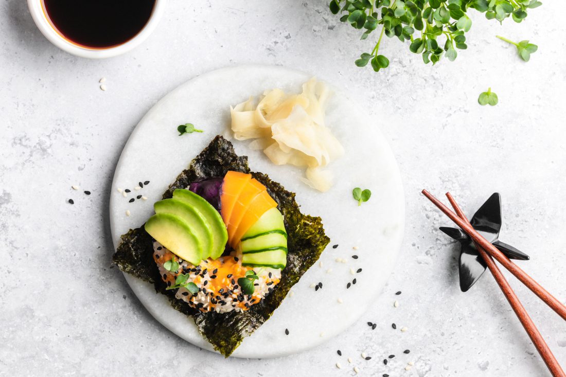 Ein bunt belegter Sushi-Donut mit Avocado, Gurke, Lachs, Rotkraut und Sesam auf einem Nori-Blatt serviert.