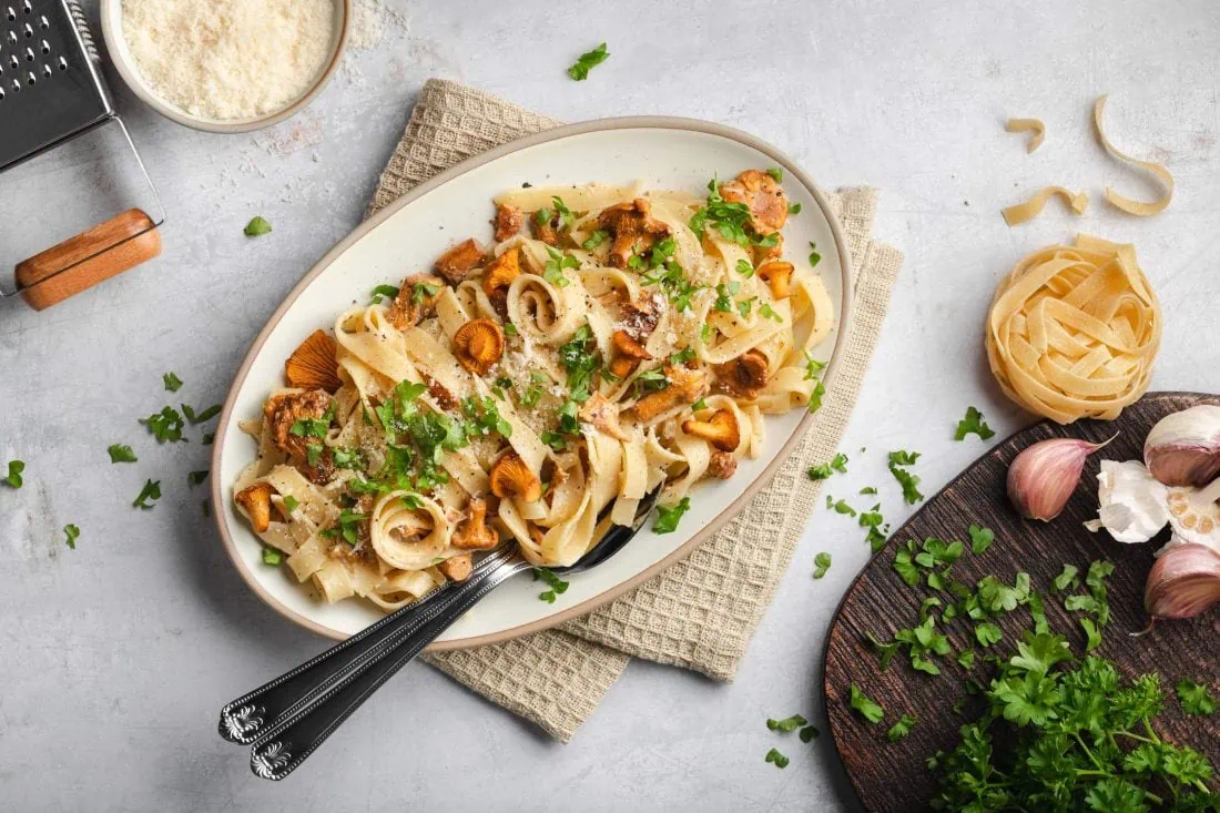 Neben den Tagliatelle mit Pfifferlingen liegt eine Käsereibe mit Parmesan und eine Schneidebrett mit weiteren Zutaten von der Zubereitung.