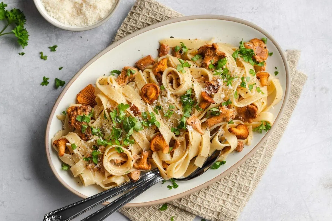Tagliatelle mit Pfifferlingen auf einem ovalen Teller bestreut mit geriebenem Parmesan.