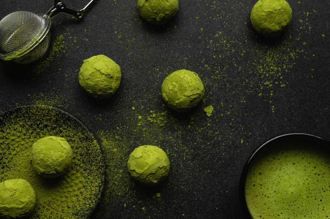 Vegane Matcha Energy Balls mit Datteln und Kokos, eine Tasse gefüllt mit Matcha Tee und ein Teesieb auf einem schwarzen Untergrund.