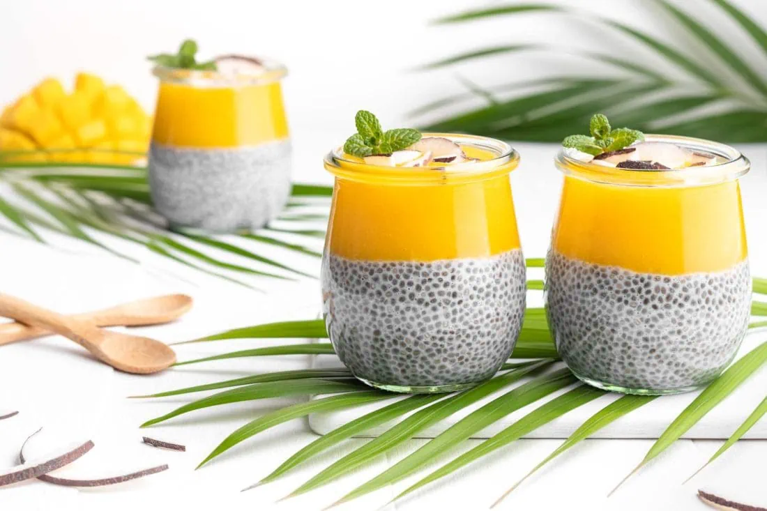 Veganer Chia Pudding mit Kokosmilch und Mangopüree in Gläsern garniert mit Kokoschips und frischer Minze.