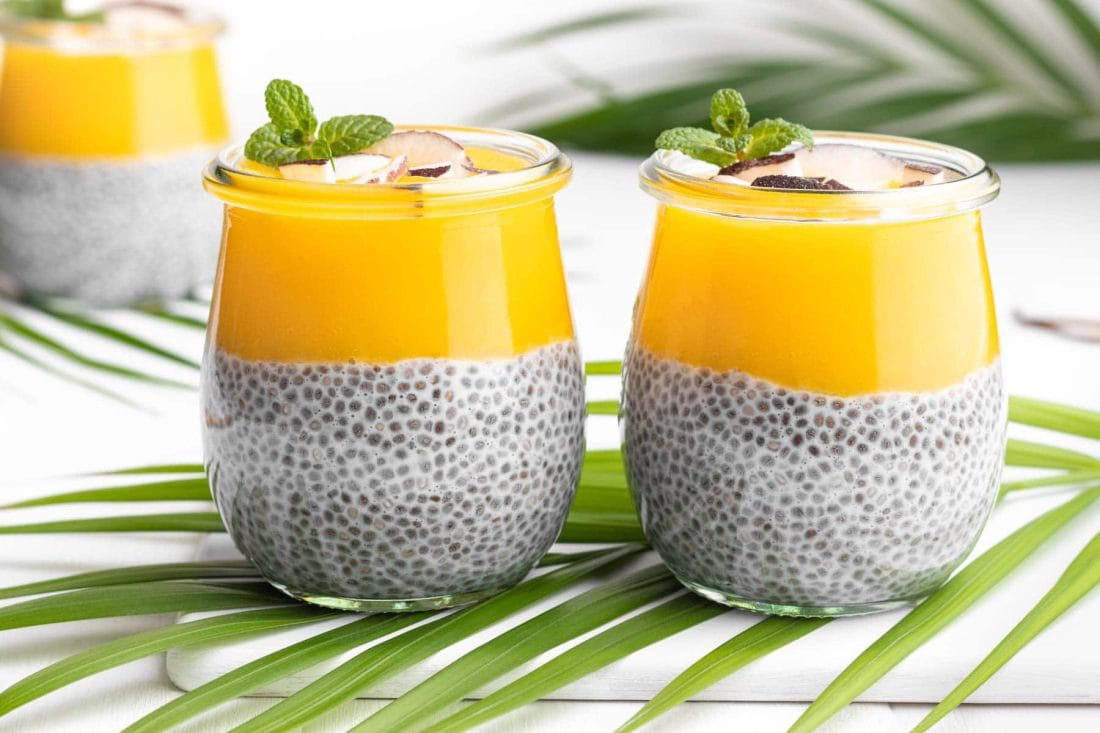 Zwei Gläser Chia Pudding mit Kokosmilch und Mango, garniert mit Kokoschips und frischer Minze, stehen nebeneinander auf einem Palmenblatt.
