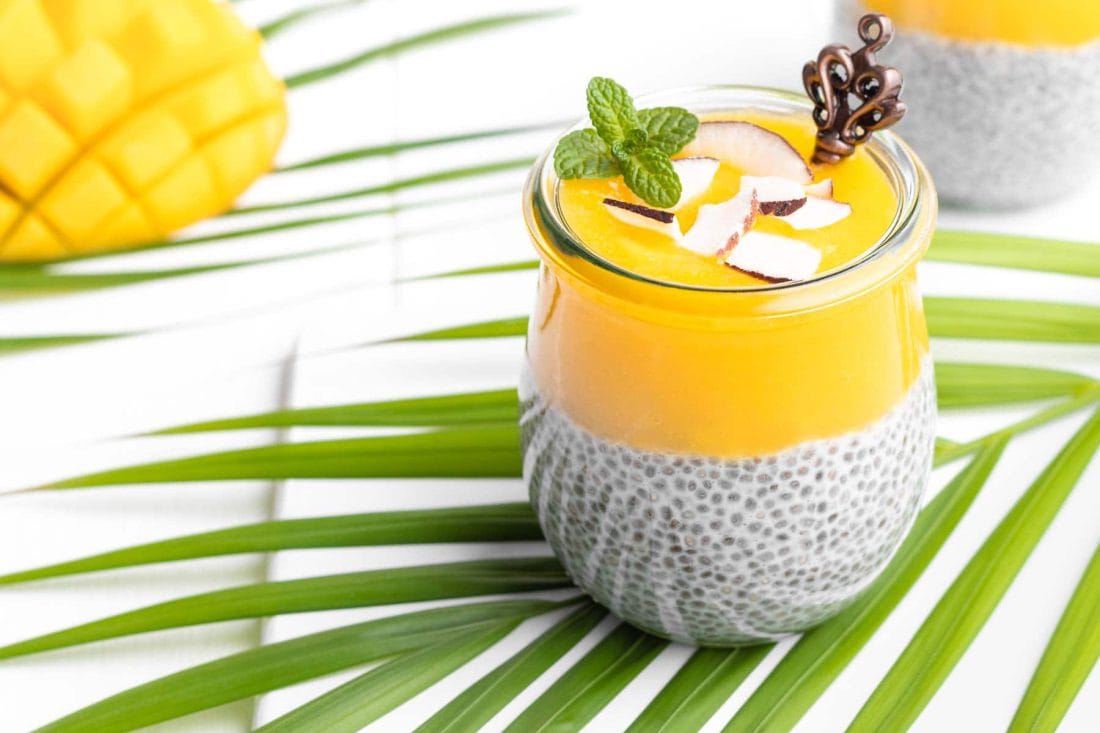 Chia Pudding mit Kokosmilch und Mango abgefüllt in einem kleinen Glas auf einem Palmenblatt.
