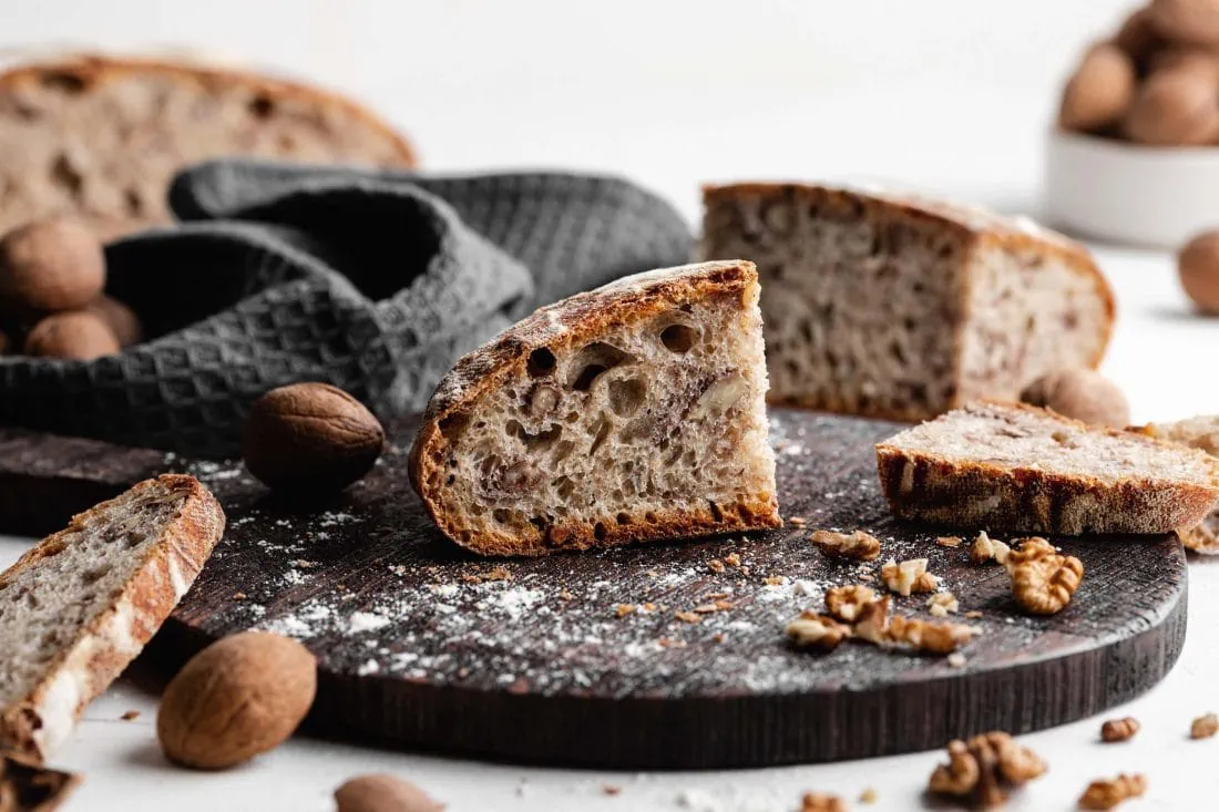 Angeschnittenes Walnussbrot mit Dinkelmehl und Sauerteig auf einem Brett. Neben dem Brot liegen Walnussstücke.