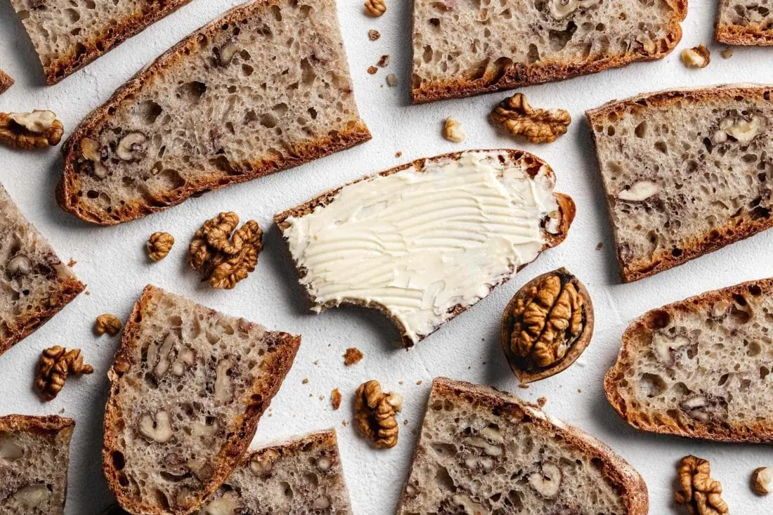 Mehrere Scheiben vom einfachen Walnussbrot mit Dinkelmehl und Sauerteig liegen auf einem weißen Untergrund. Nur die Scheibe in der Mitte ist mit Butter bestrichen.