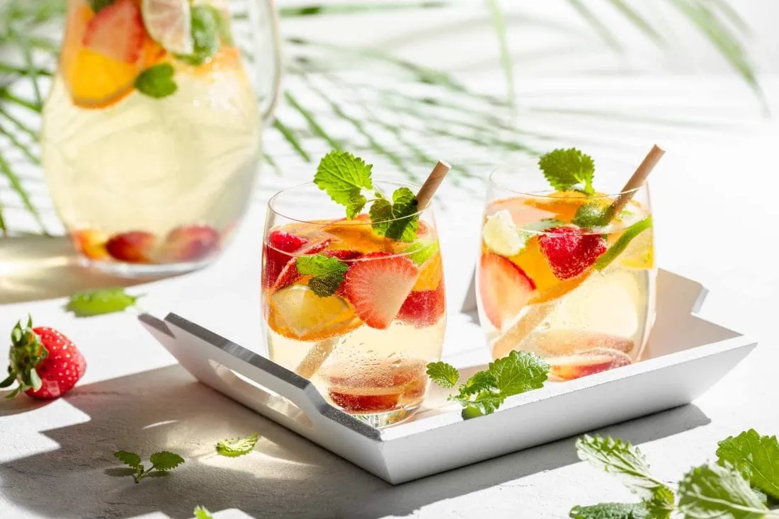 Spanische Sangria de Cava mit Sekt und Weißwein in zwei Gläsern. Darin schwimmen Erdbeeren, Trauben, Limetten, Orange und Himbeeren.