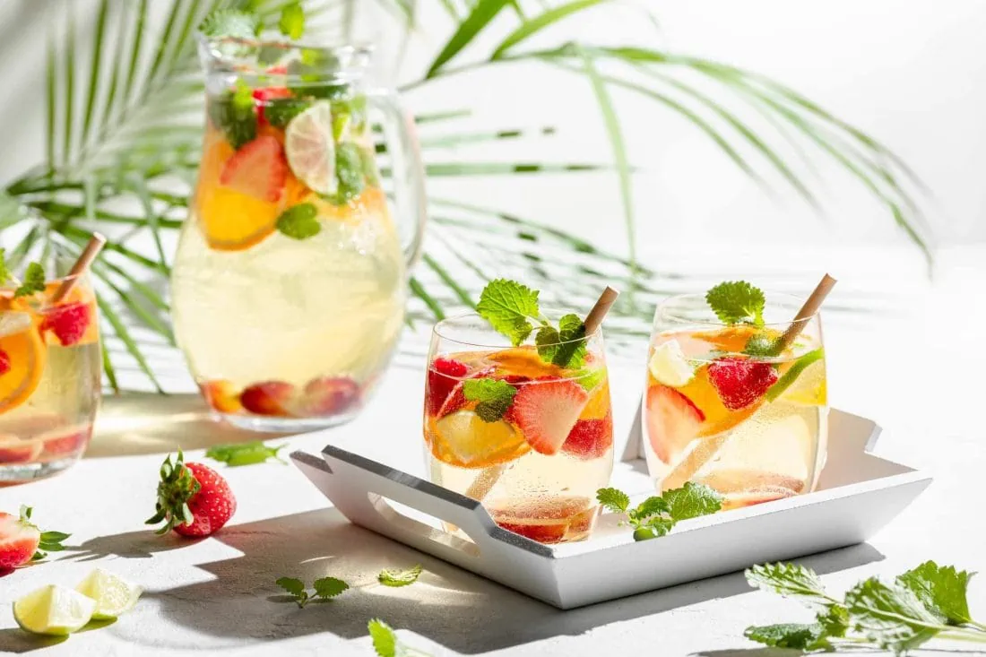 Erfrischende Sangria mit Weißwein und Sekt serviert in Gläsern serviert auf einem Tablet. Im Hintergrund steht die volle Karaffe.