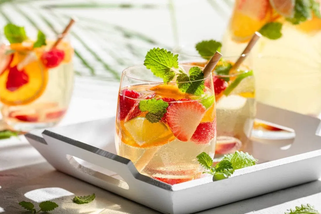 Weiße Sangria de Cava mit Sekt, Weißwein und frischen Früchten in Gläsern auf einem weißen Tablett.