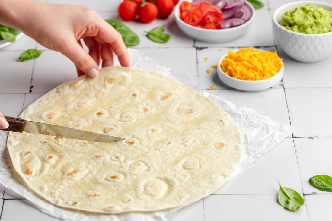 Ein Bild von einem Tortilla Wrap der vor dem Füllen und Falten bis zur Mitte eingeschnitten wird. Im Hintergrund stehen die Zutaten für die Füllung.
