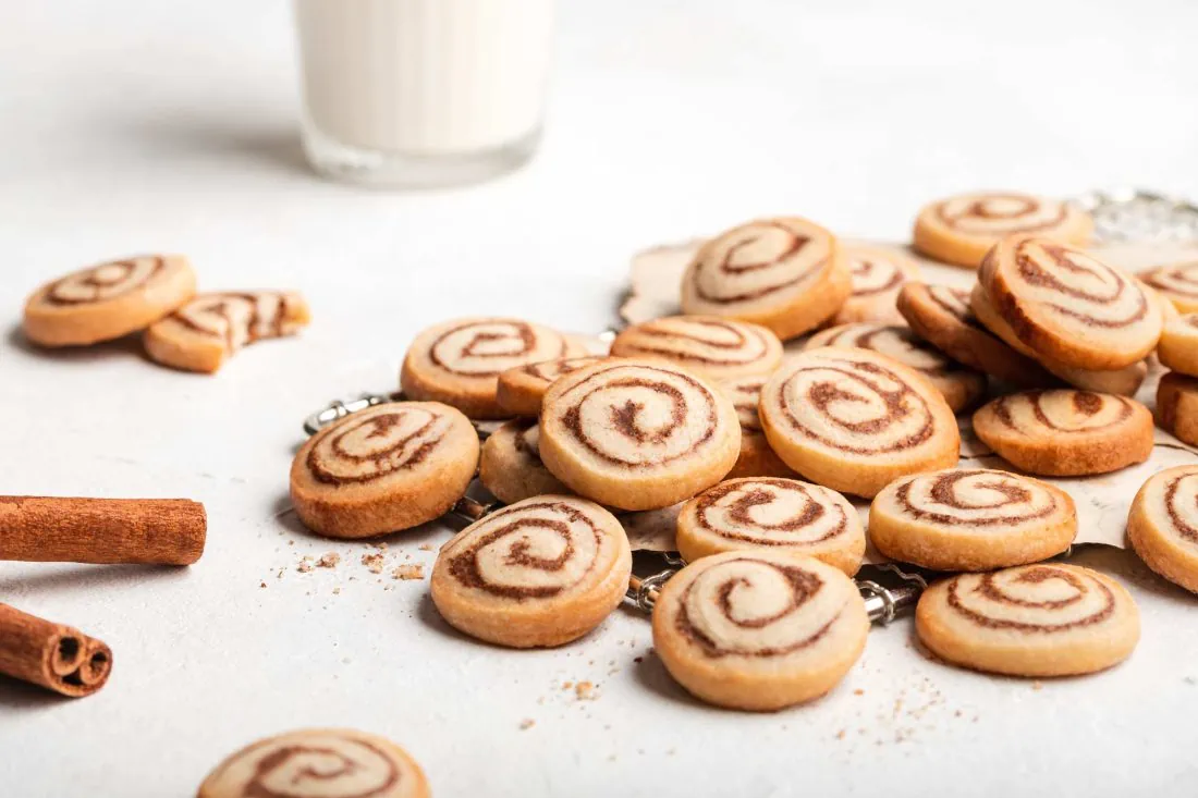 Viele kleine Zimtschnecken Plätzchen liegen aufeinander. Neben den Keksen steht ein Glas mit Milch.